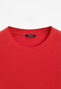 Rød bomulds t-shirt med rund hals og korte ærmer. Stoffet er glat med en ensartet tekstur; har et lille sort mærke under kraven.