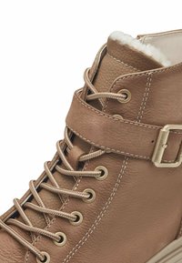 Botte de cheville en cuir marron avec lacets et une sangle à boucle. Présente une finition texturée, des œillets dorés et une doublure en fausse fourrure blanche en haut.