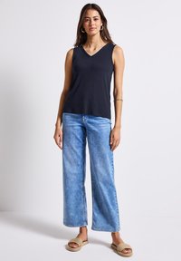 Marinblå ärmlös topp med V-ringning, kombinerad med vida ljusblå denimjeans. Modellen bär beiga vävda sandaler utan häl.