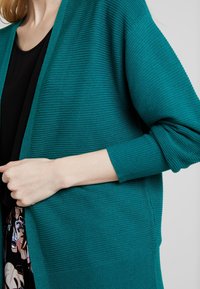 Tyrkysový pletený kardigan s ribbed texturou, otevřeným předním designem a dlouhými rukávy. Má volný střih a jemné designové detaily.