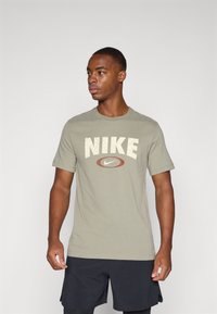 Olivengrønn t-skjorte laget av bomull, med en stor sprukken hvit "NIKE" logo og en rød oval logo sentrert under. Rund hals, korte ermer.