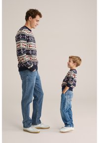 Matchande stickade tröjor med geometriska mönster i marinblått, gräddvitt och rött på en man och en pojke, kombinerade med blå jeans och vita sneakers.