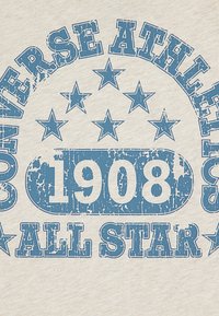Béžová textília s opotrebovaným modrým textom: "CONVERSE ATHLETIC ALL STAR" a "1908" obklopená hviezdami, v kruhovom dizajne.