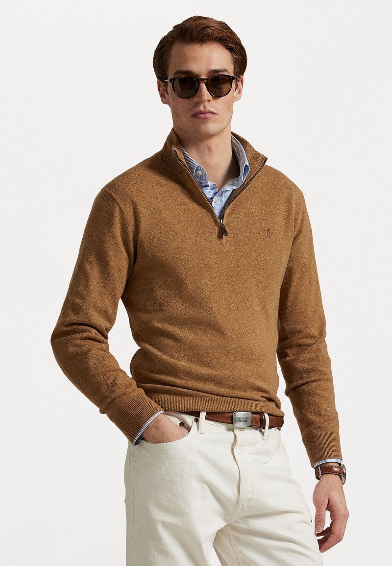 Μπλούζα φερμουάρ σε καφέ απόχρωση από υφασμάτινη ύφανση, φορεμένη πάνω από μπλε ριγέ πουκάμισο, με κρεμ παντελόνι και καφέ ζώνη. Συμπεριλαμβάνονται μαύρα γυαλιά ηλίου.