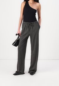 Bodysuit noir à une épaule associé à un pantalon large à rayures verticales noires et grises, avec une taille élastique et un cordon de serrage. Sac à main et chaussures noires.