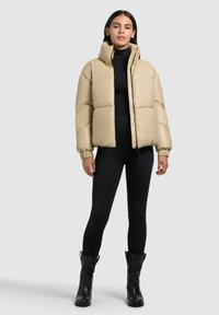 Beige puffertakó magas gallérral, cipzárral és elasztikus mandzsettákkal. Fekete felsőn viselve, fekete leggings-szel és bokacsizmákkal párosítva.