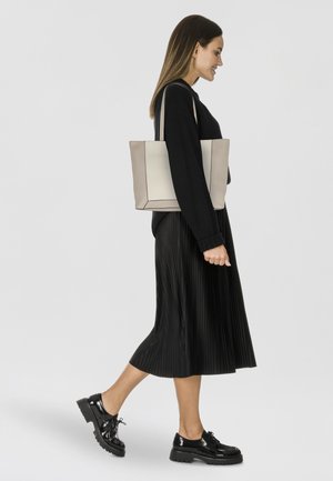 Sac fourre-tout beige clair avec des coutures noires, en forme rectangulaire, en matériau synthétique ; le modèle porte une jupe noire plissée et des chaussures brillantes.