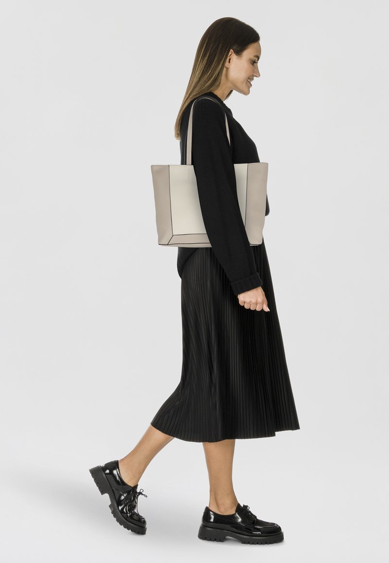 Bolso de mano en beige claro con costuras negras de acento, forma rectangular, material sintético; el modelo lleva una falda negra plisada y zapatos brillantes.