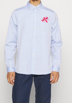 Personne portant une chemise bleu clair boutonnée avec une fleur rouge et une broderie visage souriant sur la poitrine, associée à un pantalon bleu foncé.