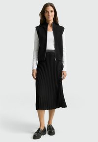 Gilet en tricot côtelé noir avec fermeture éclair, porté sur une chemise blanche à manches longues, associé à une jupe plissée noire et des mocassins noirs épais.