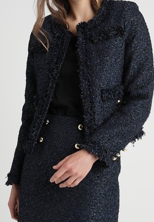 Frau trägt eine dunkelblaue, strukturierte Tweedjacke und passenden Rock mit goldenen Knöpfen und posiert mit der Hand nahe ihrer Taille.