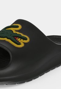 Μαύρο σανδάλι slip-on με υφασμένο πέλμα και κεντητό πράσινο λογότυπο κροκοδείλου με κίτρινο περίγραμμα στην κορυφή.