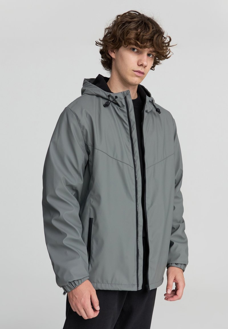 Giacca impermeabile grigia con zip frontale, cappuccio regolabile e polsini elasticizzati. Presenta due tasche laterali e una texture liscia e leggera.