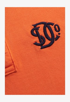 Orange fabric with dark blue embroidered stylized letters forming a logo, next to a folded fabric edge.