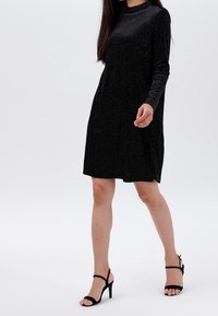 Černé dlouhé šaty s puntíkovým vzorem, s vysokým výstřihem a volným střihem. Nosí se s černými sandály na vysokém podpatku.