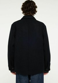 Svart ulljacka med en strukturerad krage, översized passform och knäppta manschetter. Slät textur med en ren design på bakpanelen.