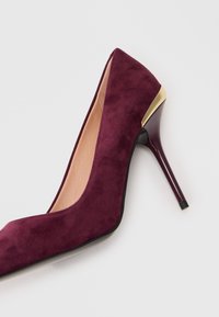 Μπορντώ σουέντ παπούτσι με στιλέτο τακούνι και χρυσή λεπτομέρεια πίσω, με καφέ εσωτερική επένδυση, φωτογραφημένο σε λευκό φόντο.