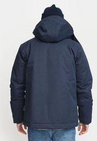 Navyblauwe geïsoleerde jas met een capuchon, voorzien van een zachte textuur en een iets langere achterzoom. Inclusief elastische manchetten voor een goede pasvorm.