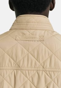 Beige gesteppte Jacke aus strukturiertem Stoff, mit Stehkragen und gesteppten Rautenmuster. Gesticktes Markenlabel auf der Rückseite.