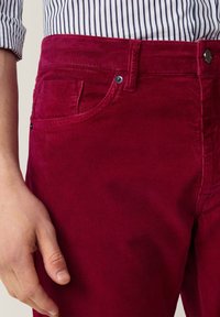 Rote Cordhosen im Slim-Fit-Stil mit fünf Taschen und subtiler Textur. Metallknopf und Nieten am Bund sichtbar.