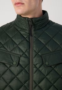 Gilet matelassé vert avec un col haut, une fermeture éclair à l'avant et deux poches poitrine avec fermetures à pression. Texture lisse et brillante avec un motif en diamant.