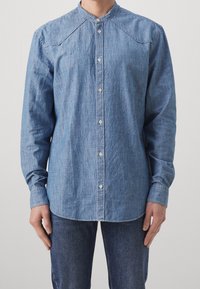 Ljusblå denimskjorta med ståkrage, långa ärmar, knappstängning och subtil textur. Har axelsömmar och knappdetaljer.