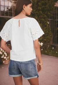 Blouse blanche avec des détails en œillets et une ouverture en forme de clé au dos, associée à un short en denim retroussé avec deux poches arrières.