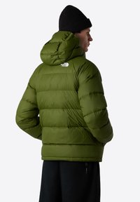 Grön puffjacka med huva, med en quiltad design och elastiska manschetter. Logotyp på baksidan, bärs över svarta fleecebyxor.