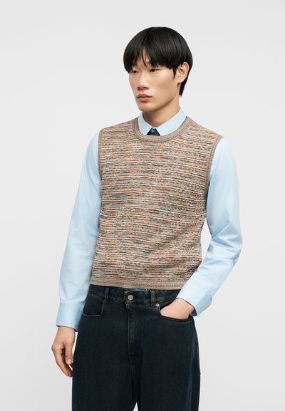 Homme portant un gilet tricoté multicolore sur une chemise bleu clair à boutons et un jean noir, debout avec une main dans la poche.