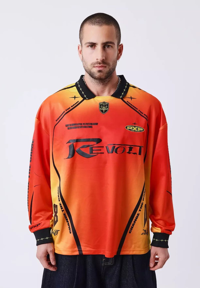 Homme portant un t-shirt à manches longues dégradé rouge-orange avec le texte noir "REVOLT" et un col noir, debout devant un fond clair uni.