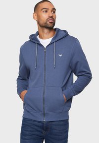 Blaues Zip-Hoodie mit einer Kordelzugkapuze, Fronttaschen und einem kleinen Logo auf der Brust. Hergestellt aus weichem Stoff mit gerippten Bündchen.