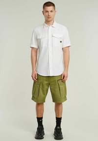 Camisa blanca de manga corta con botones y dos bolsillos frontales, combinada con pantalones cortos de cargo verde olivo que tienen bolsillos laterales. Zapatillas negras y calcetines.