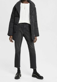 Schwarze Daunenjacke mit großem Kragen, getragen über einer weißen Bluse. Kombiniert mit hochgeschnittenen schwarzen Jeans und glänzenden schwarzen Schuhen.