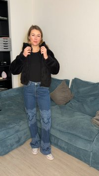 Kvinne som justerer svart pelsjakke, iført svart topp, blå mønstrete jeans og hvite joggesko, stående ved siden av en grønnblå sofa med puter.