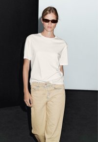 T-shirt bianco in cotone abbinato a jeans larghi beige chiaro. Il modello indossa occhiali da sole scuri, in piedi contro uno sfondo contrastante nero e chiaro.