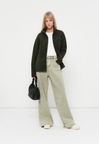 Giacca in pile verde con zip, t-shirt bianca, jeans larghi verde chiaro e borsa nera. Caratteristiche: vestibilità rilassata e tessuto testurizzato.