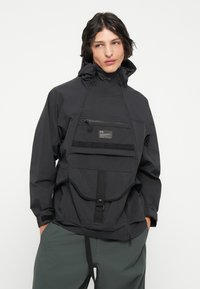 Sort pullover-jakke med høj krave, justerbar hætte, frontlomme med lynlås og stor frontlomme med spænde-lukning. Letvægtsstof.
