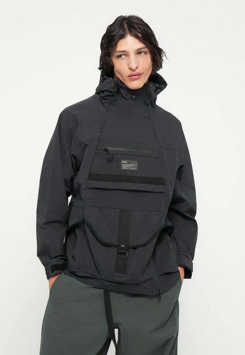 Giacca a pullover nera con colletto alto, cappuccio regolabile, tasca frontale con zip e grande tasca frontale con chiusura a fibbia. Tessuto leggero.