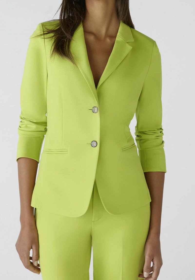 Blazer vert lime en tissu lisse, avec des revers crantés, des manches longues, deux boutons et des poches latérales, associé à un pantalon assorti.