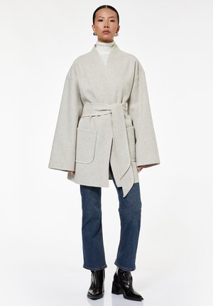 Vrouw draagt lichtgrijze wollen jas met ceintuur over een witte coltrui, blauwe spijkerbroek en zwarte enkellaarsjes met hak, staand tegen een witte achtergrond.