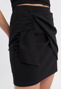 Schwarzer Rock aus strukturiertem Stoff mit einem geknoteten Detail vorne, taillierter Silhouette und einem elastischen Bund für zusätzlichen Komfort.