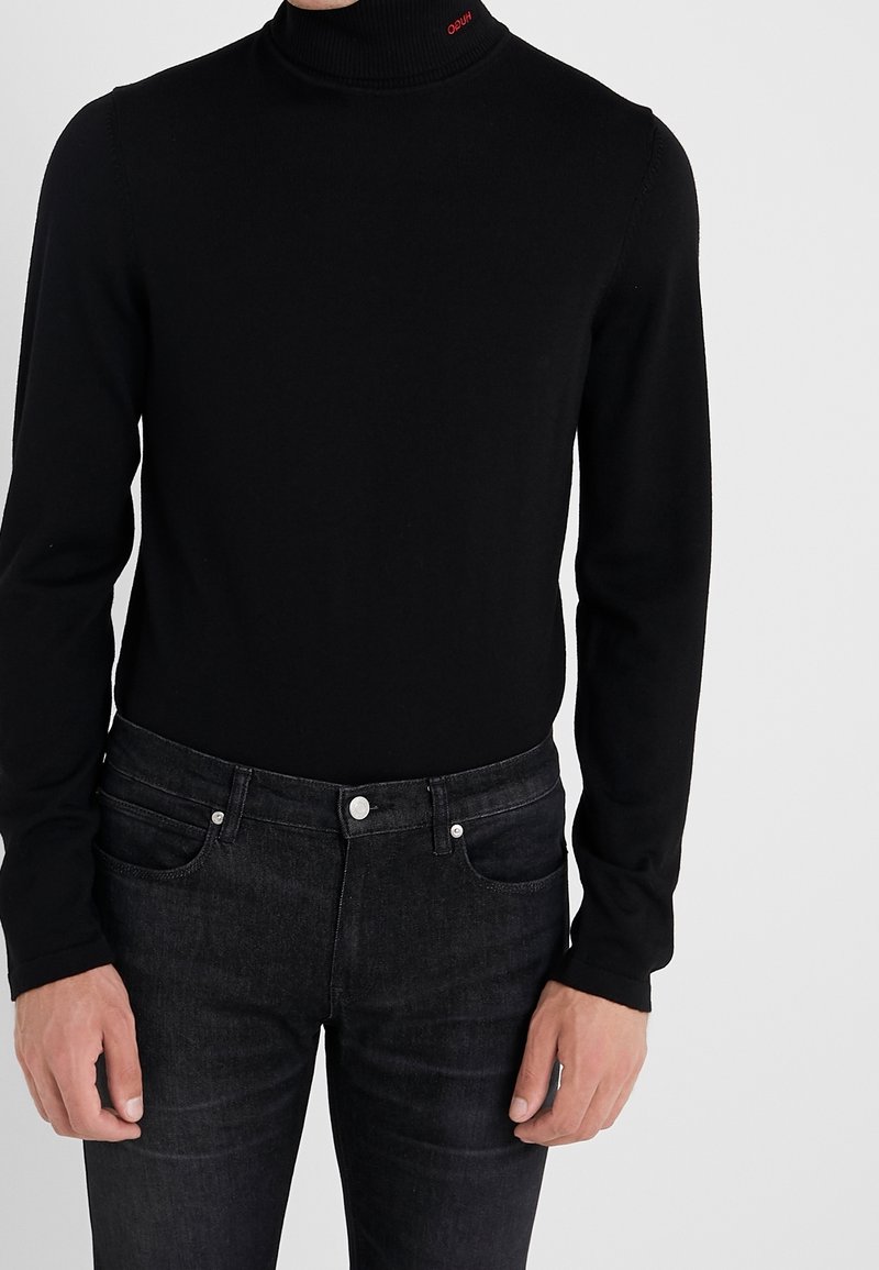 Homme portant un pull noir ajusté à col roulé et un jean slim gris foncé, debout avec les mains relâchées le long du corps sur un fond blanc.