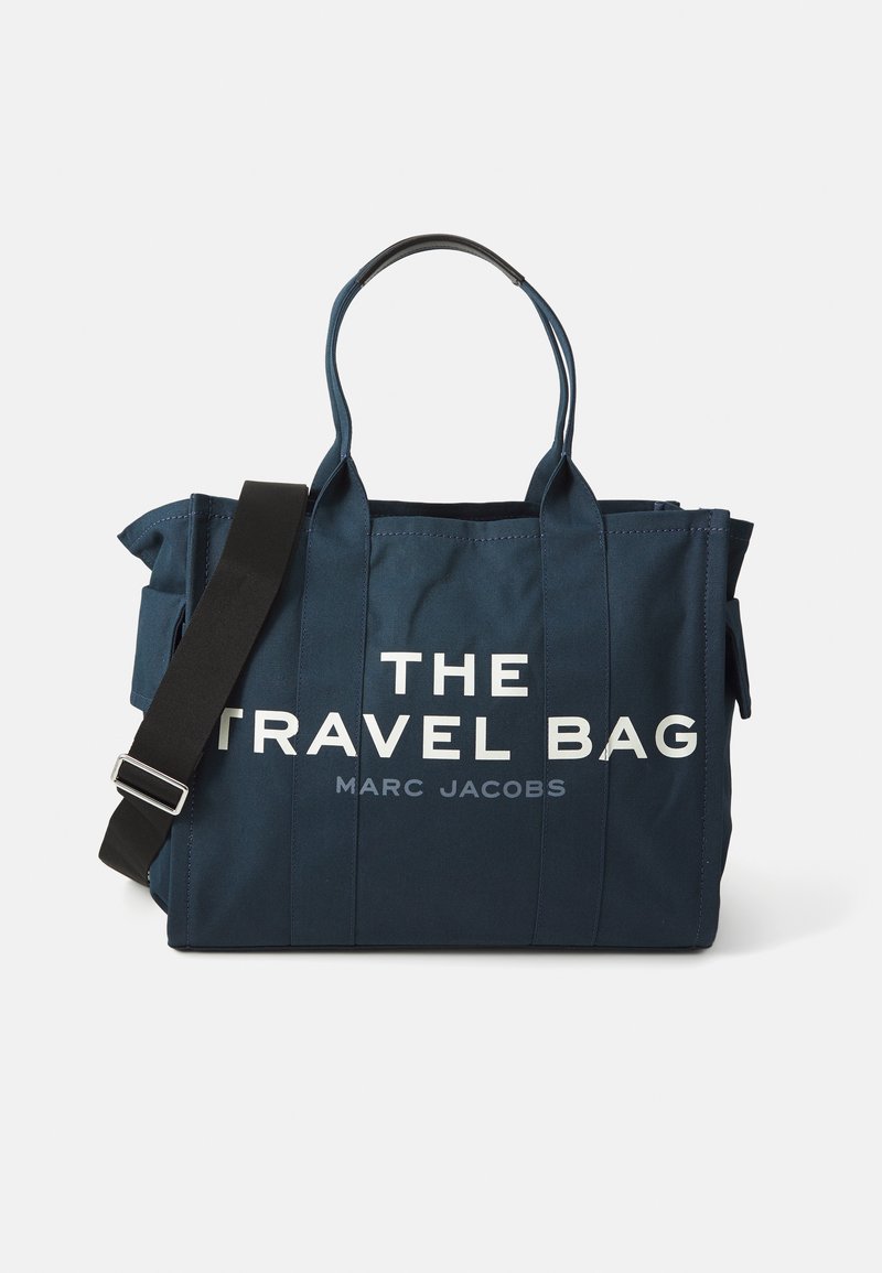 Saco de viagem em tecido azul marinho com duas alças e uma alça removível, apresentando o texto em branco em destaque: "O SACO DE VIAGEM MARC JACOBS."