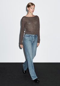 Hnědý pletený svetr se širokým výstřihem v kombinaci se světle modrými kalhotami s přímým střihem. Modelka má na nohou černé dřeváky. Jednoduché pozadí.