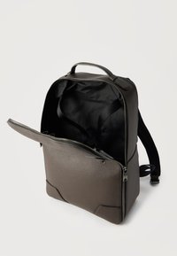 Καφέ δερμάτινο σακίδιο πλάτης με μαύρο εσωτερικό και μερικώς ανοιχτό φερμουάρ μπροστά σε λευκό φόντο.