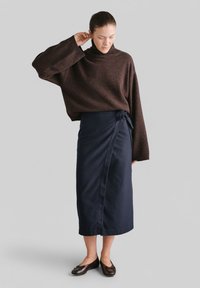 Brązowy sweter z golfem, dziergany, z szerokimi rękawami, w zestawieniu z ciemnoniebieską spódnicą kopertową. Modelka nosi brązowe baleriny, stojąc na szarym tle.