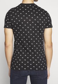 Czarny t-shirt z krótkim rękawem z nadrukiem białego logo CK na całej powierzchni. Materiał jest gładki i dopasowany, z okrągłym dekoltem i standardowym wykończeniem dołu.