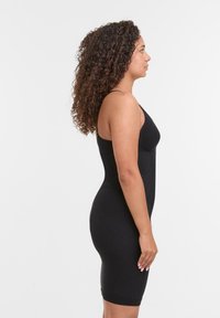 Donna con capelli ricci, vestita con un abito nero aderente senza maniche, in piedi di profilo su uno sfondo bianco uniforme.