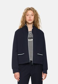 Navy bomber jacket made from soft fabric, featuring a high collar, white zipper, striped pockets, and a relaxed fit, worn over a striped shirt.