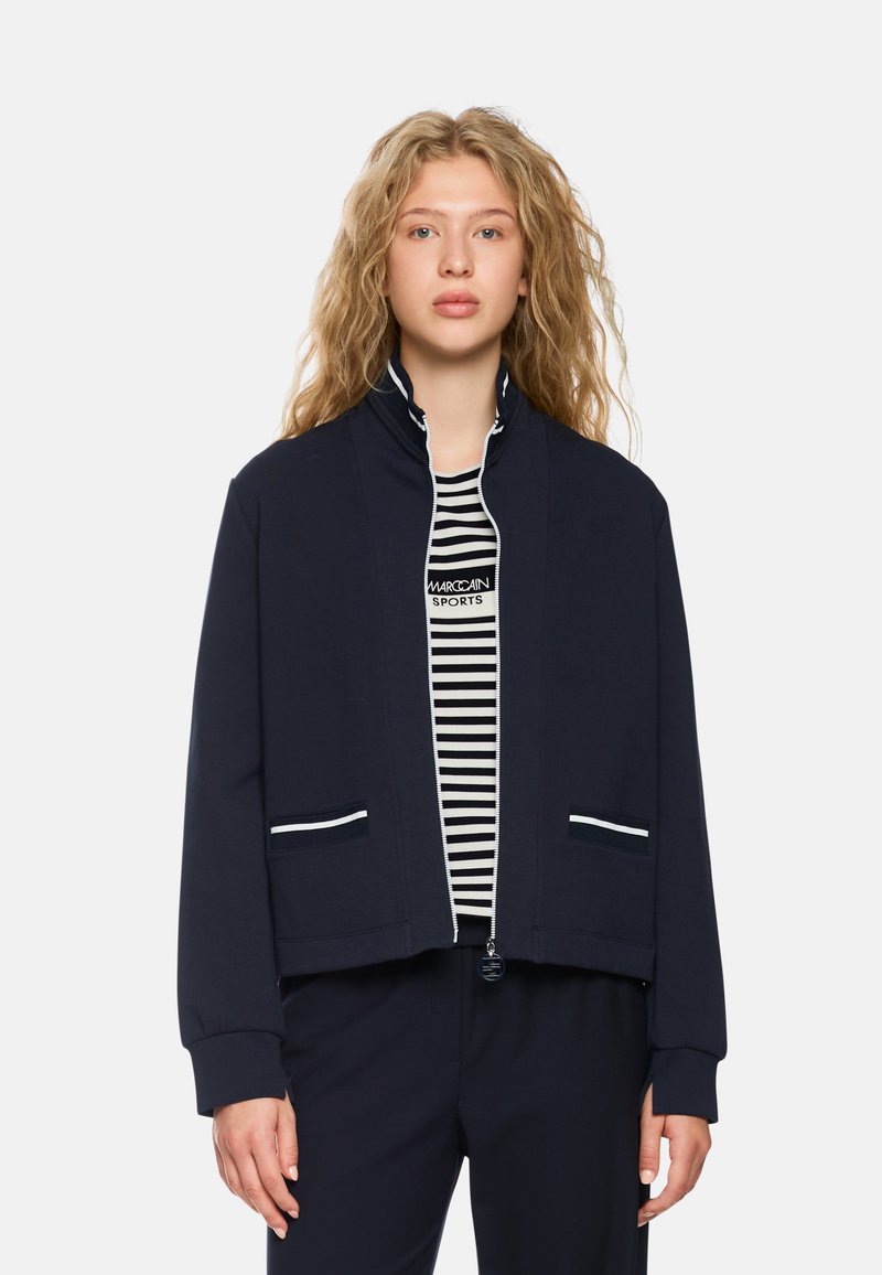 Navy bomber jacket made from soft fabric, featuring a high collar, white zipper, striped pockets, and a relaxed fit, worn over a striped shirt.
